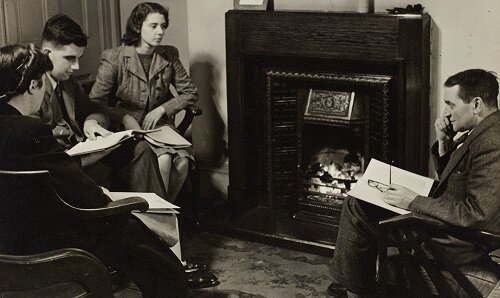 Group of male and female students sat near a fire place listening to their tutor