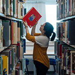 Person reaching for a book on a high shelf between library aisles.