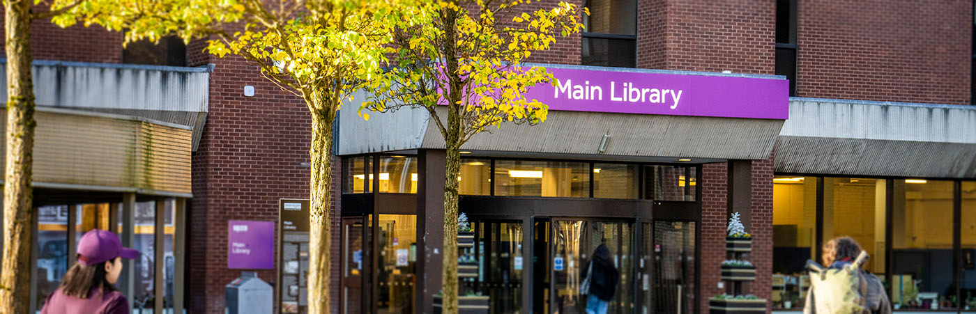 Entrance to the Main Library at The University of Manchester, with a purple sign above the doors and people walking nearby.