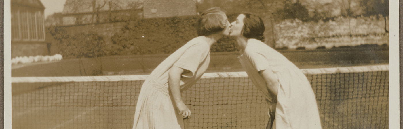 Photograph of two women kissing in front of a net on a tennis court. They are both holding rackets