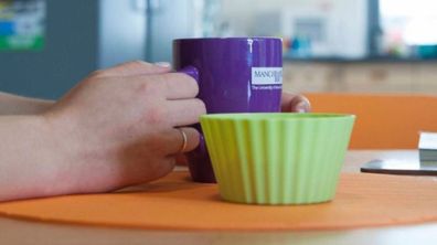 A purple University of Manchester mug held by someone sat at a table in a kitchen.