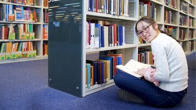 A student holds a book while sitting on the floor in front of a shelf of books