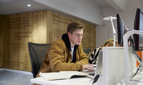 Student sits at a computer in one of the cluster in AGLC with a textbook.