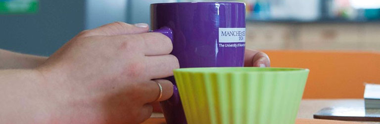 Hands hold a University of Manchester mug filled with a hot drink.